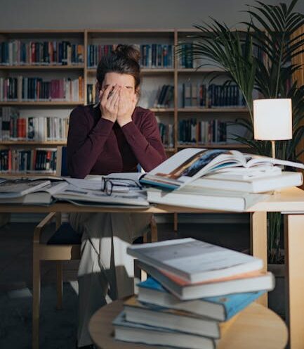 stressed out purpose driven professional A woman looks stressed while studying with books in a dimly lit library during nighttime.